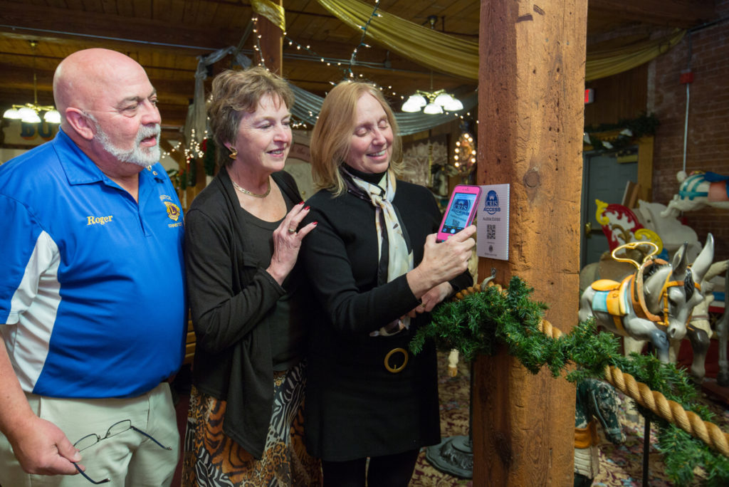 man in blue shirt with two women wearing black tops viewing the use of a smartphone to scan a QR code for the carousel exhibit