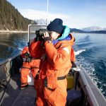 a person in an orange insulated jumpsuit with a navy hat looking through binoculars while standing on a boat in water surrounded by snow capped mountains