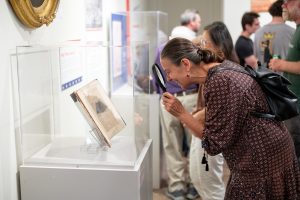 a women smiling and holding a magnifying glass to look at an exhibit housed in a glass case