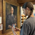 a young girl standing in front of a portrait of a man using her phone to listen to the audio description
