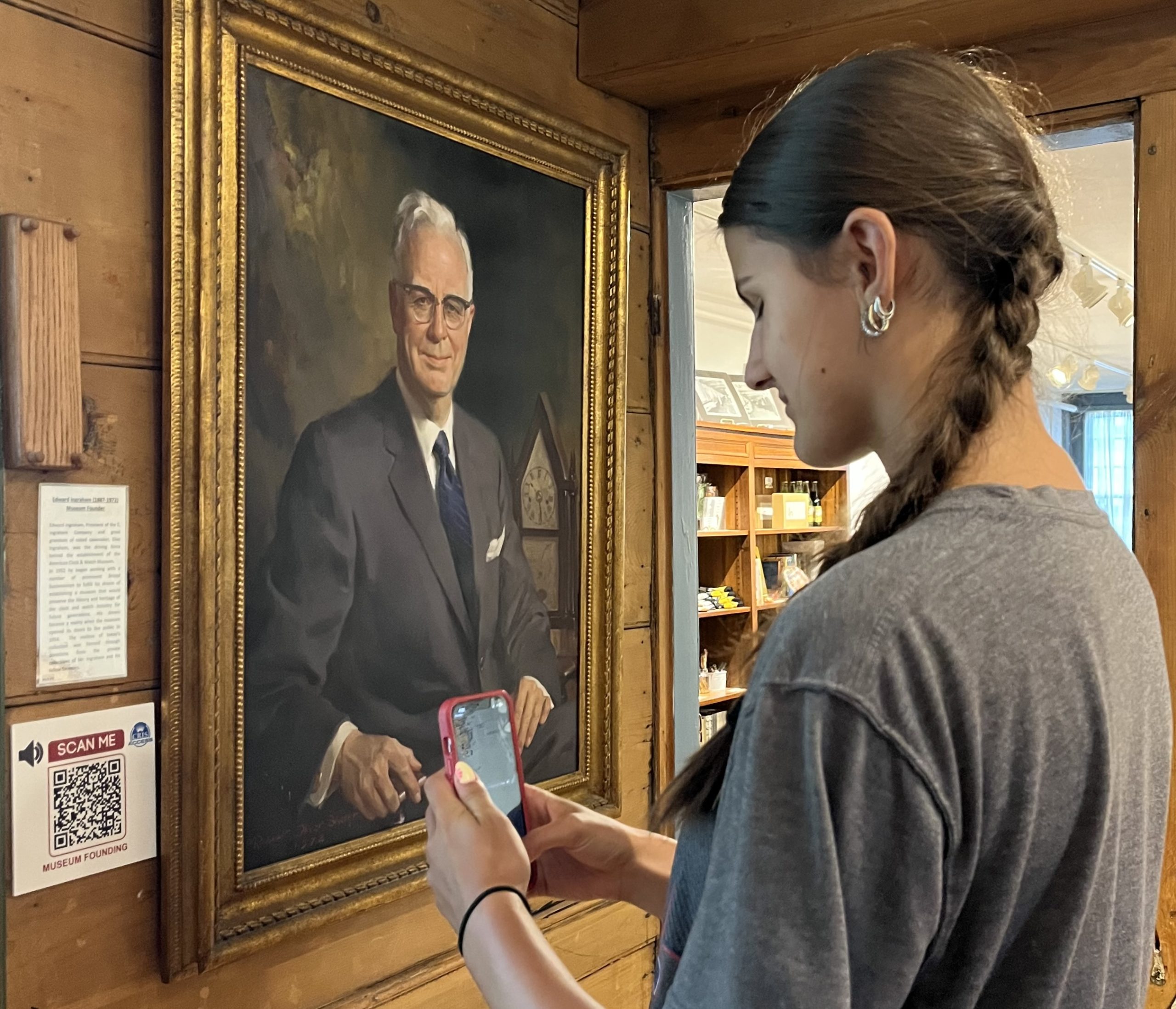 a young girl standing in front of a portrait of a man using her phone to listen to the audio description