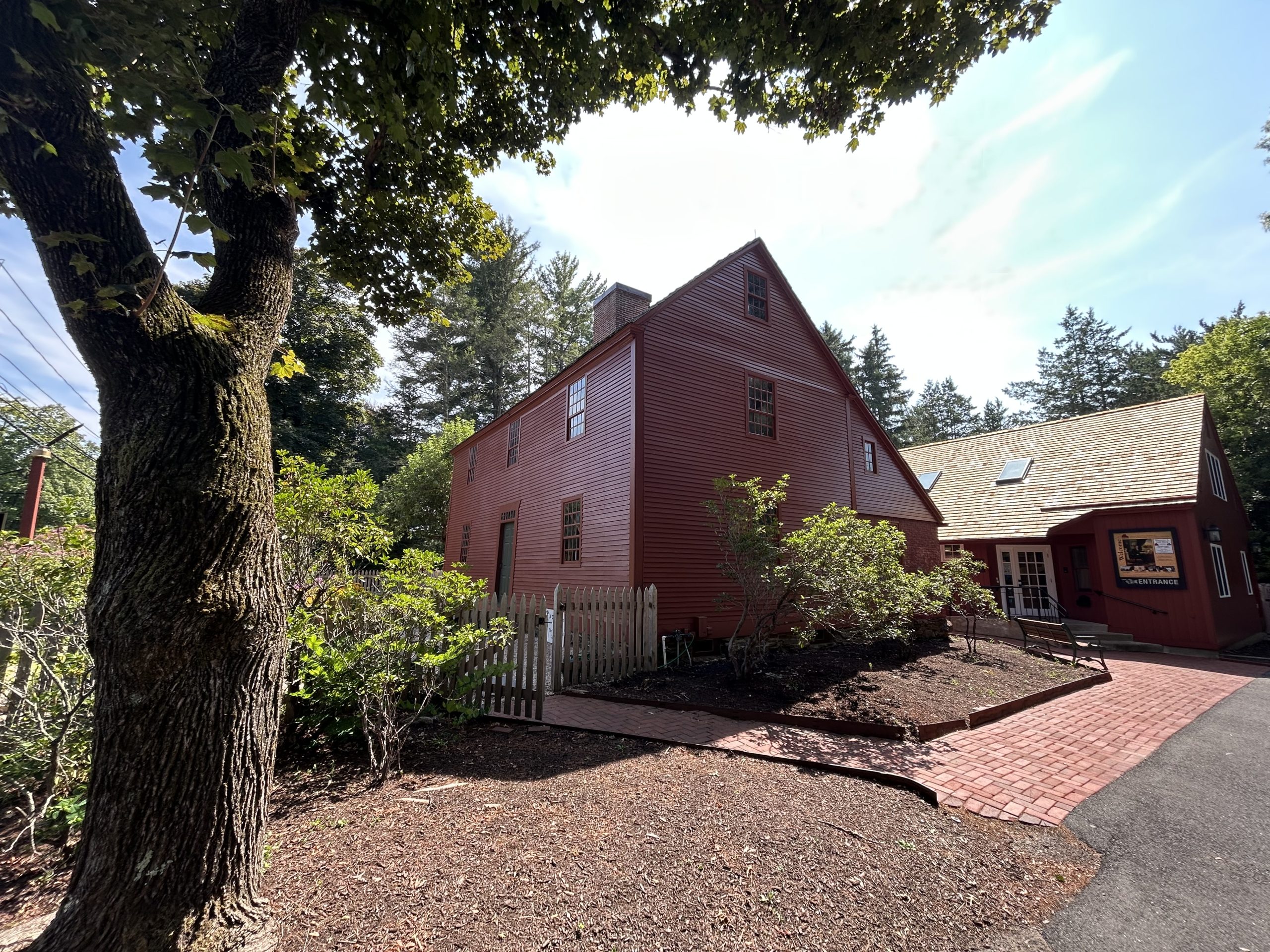 Angled view of a historic red colonial house with a steep roof and brick chimney, next to a modern visitor entrance building, with trees, shrubs, and a brick walkway in the foreground.