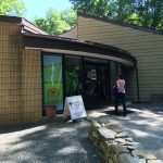 Front entrance of a museum building with glass doors, a curved roof, and a visitor walking toward the entrance past a sign and paved walkway.