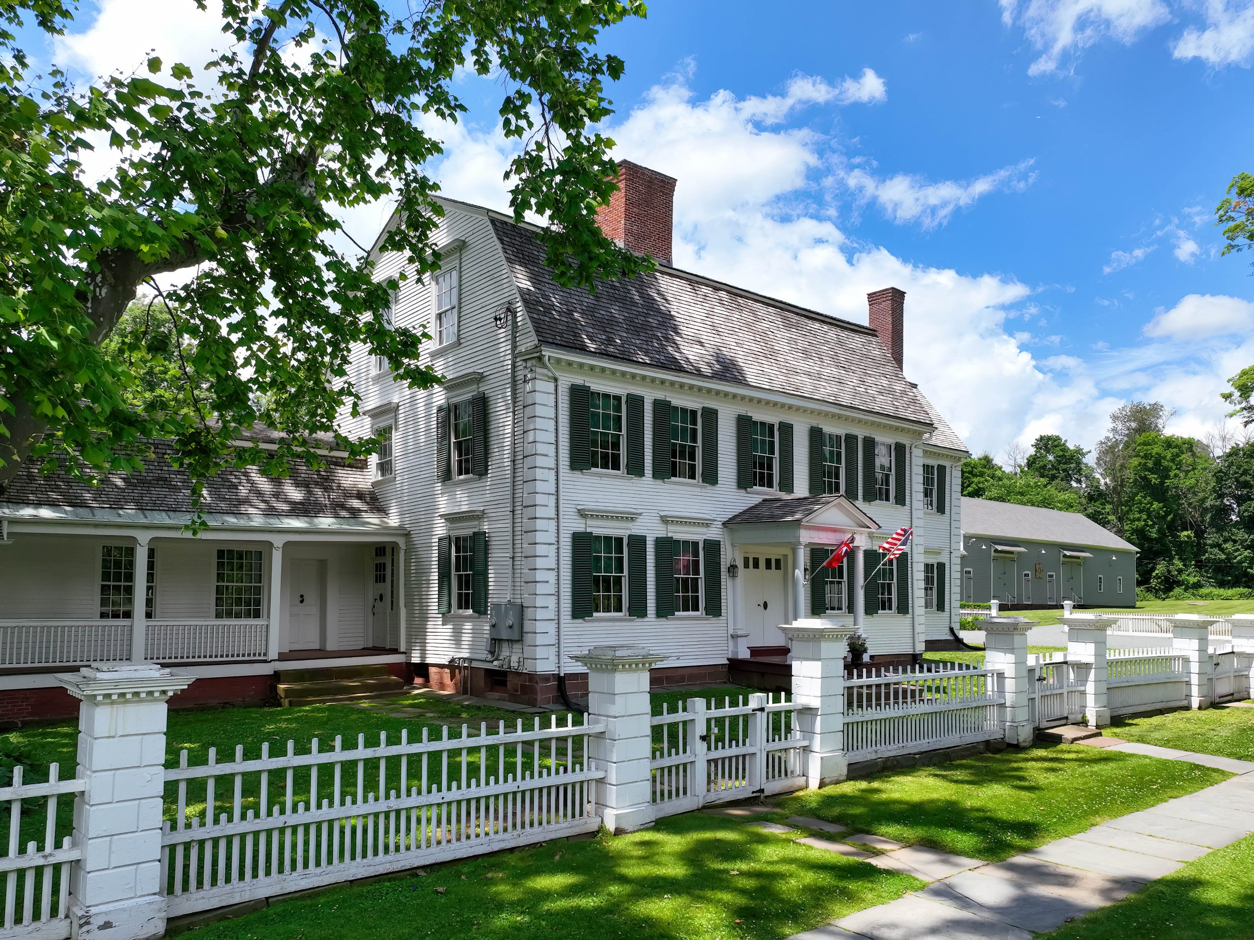 Exterior view of a large, historic two-story white Colonial-style home with black shutters, red brick chimneys, and a covered front porch. A white picket fence surrounds the property, with two American flags displayed near the main entrance. Mature shade trees frame the house under a bright blue sky with scattered clouds, and a gray outbuilding is visible in the background.