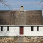 Beaumont House “Historic single-story white wooden house with a steep shingled roof, central stone chimney, red front door, and symmetrical windows, viewed behind a low stone wall.”