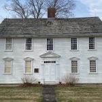 Governor Jonathan Trumbull House “Front view of the historic two-story white Governor Jonathan Trumbull House, featuring symmetrical multi-pane windows, a central doorway with classical detailing, a brick chimney, and mature trees.”