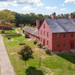 Aerial view of a historic homestead featuring a large two-story red wooden building with multiple brick chimneys, surrounded by smaller red and wood buildings, stone walls, grassy grounds, and tree-lined paths under a blue sky.