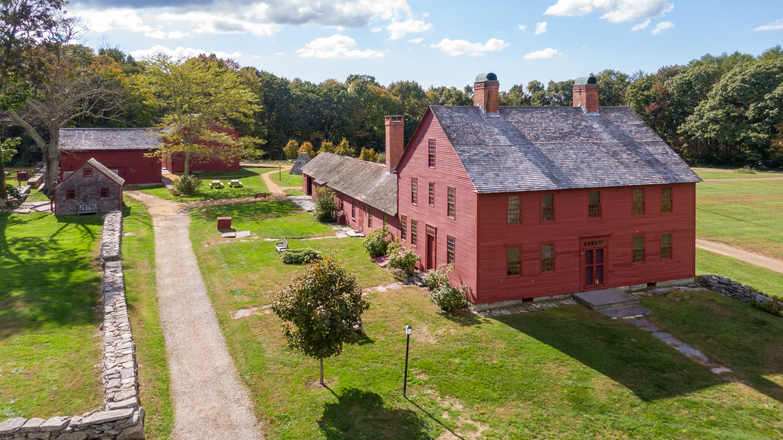 Aerial view of a historic homestead featuring a large two-story red wooden building with multiple brick chimneys, surrounded by smaller red and wood buildings, stone walls, grassy grounds, and tree-lined paths under a blue sky.