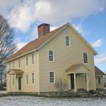 Jonathan Trumbull Jr. House “Historic two-story pale yellow wooden house with a steep gabled roof, brick chimney, multi-pane windows, and covered entrances, surrounded by trees with a light dusting of snow on the ground.”