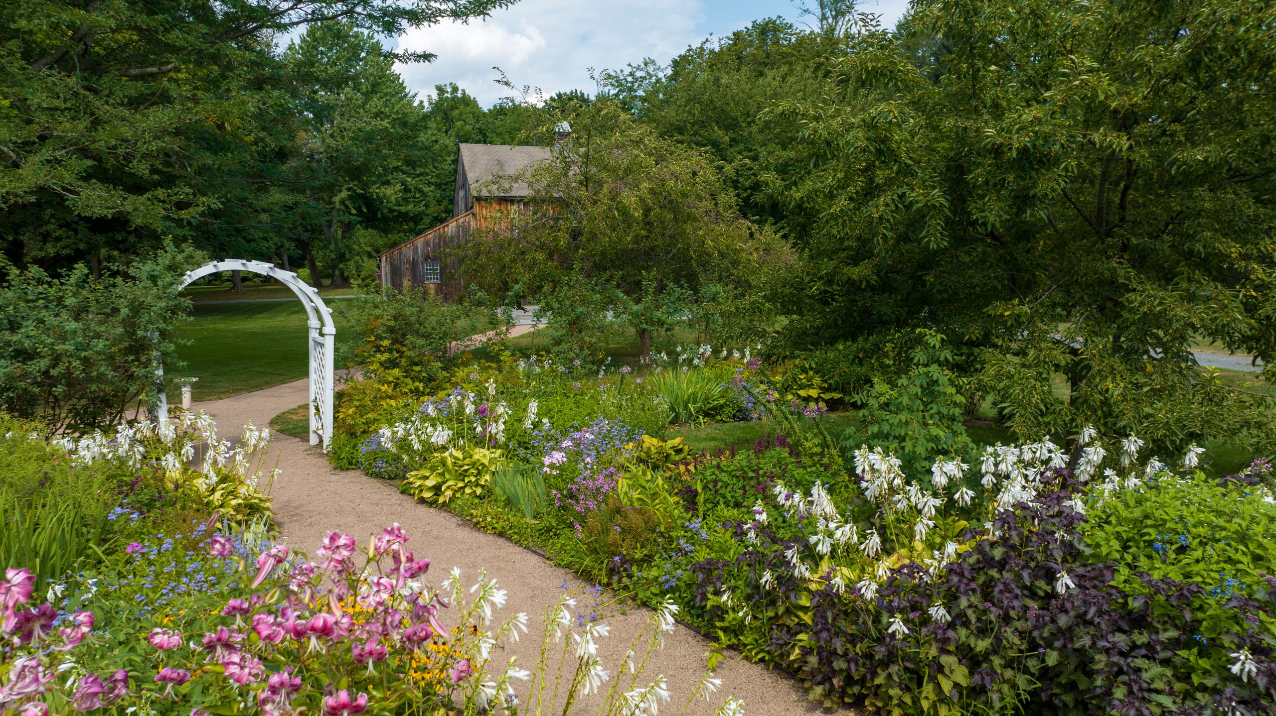 Colorful garden at the Webb Deane Stevens Museum featuring winding paths bordered by pink, purple, blue, and white flowers and lush greenery. A white garden arbor and historic wooden building are visible among mature trees.